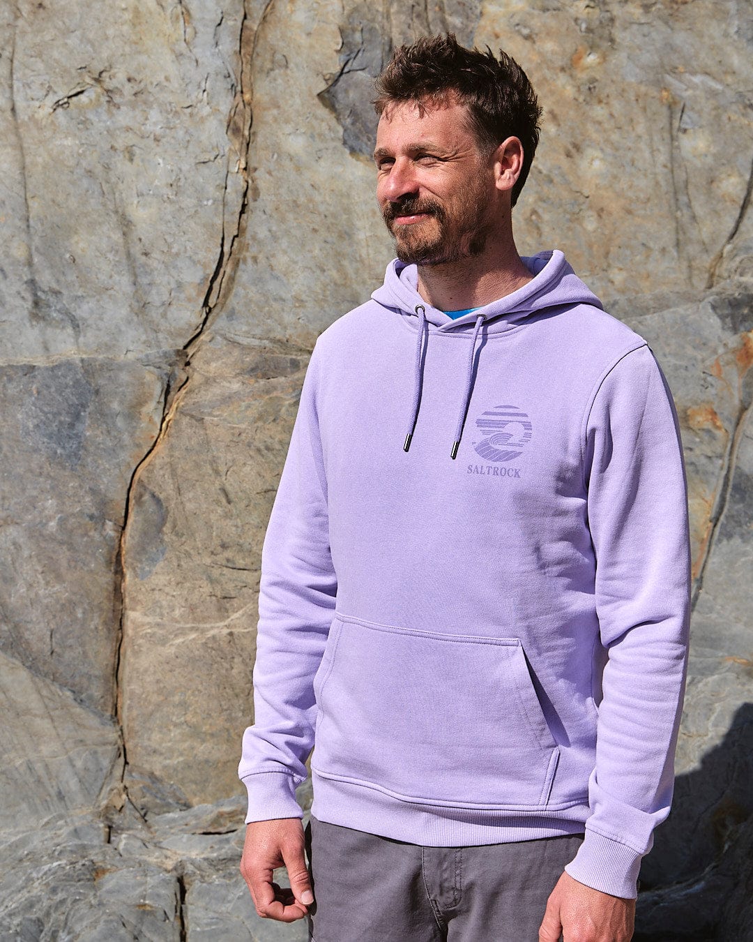 A man wearing a Saltrock Atlantic - Men's Pop Hoodie - Purple standing next to a rock wall.