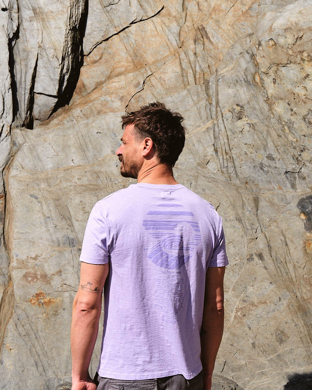 A man wearing a Saltrock Atlantic - Mens Short Sleeve T-Shirt in Purple standing in front of a rock.