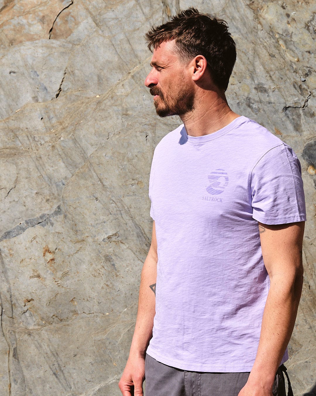 A man wearing an Atlantic - Mens Short Sleeve T-Shirt - Purple by Saltrock, standing next to a rock wall.