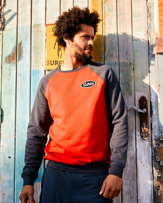 Man in a Saltrock red and gray sweatshirt with raglan contrast sleeves standing by a weathered wooden wall with a surf sign, looking to the side.