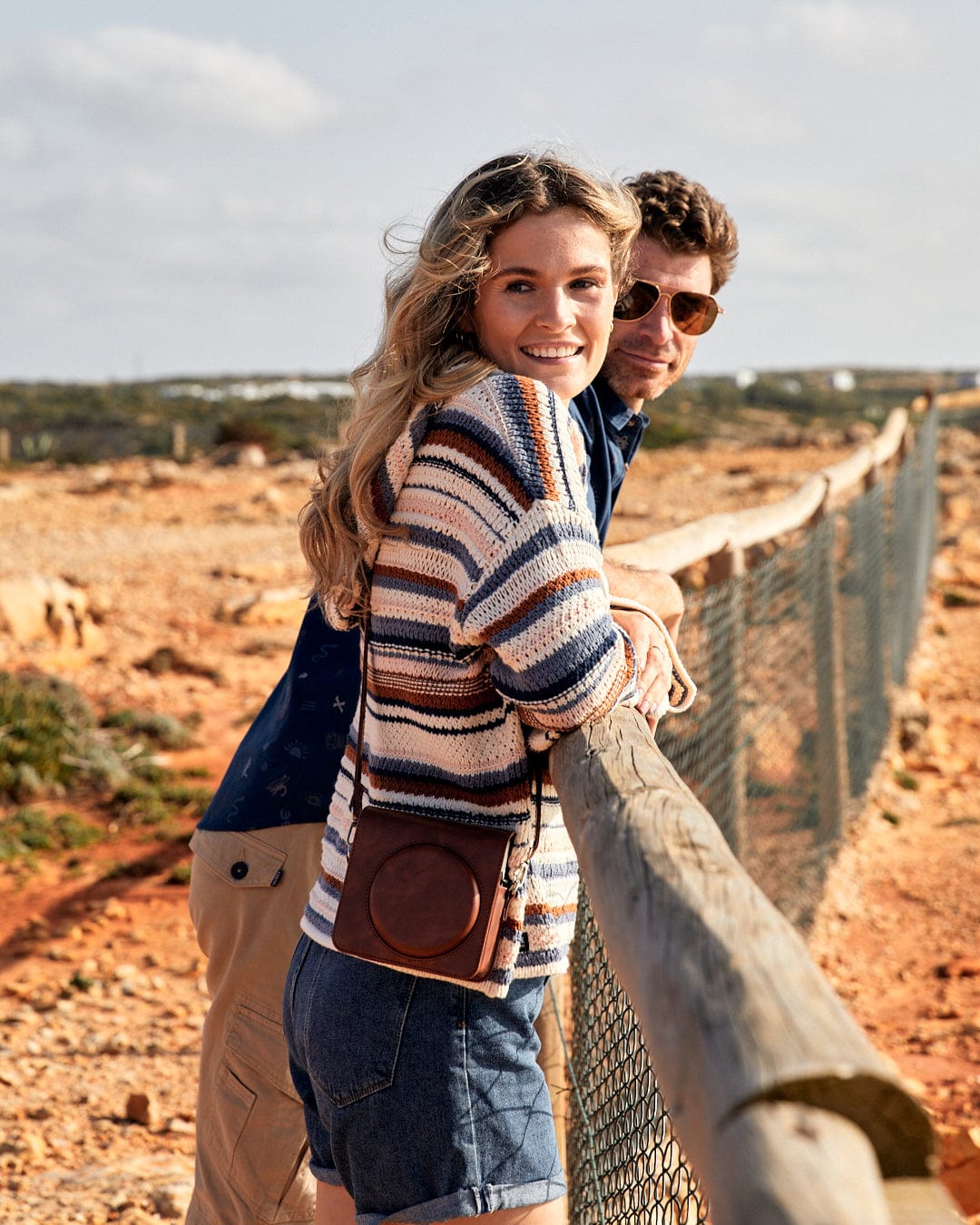 A man and woman leaning against a fence in the desert, with draw cords, wearing Saltrock's Aubrey Pop - Womens Hooded Crochet Knit in Cream.