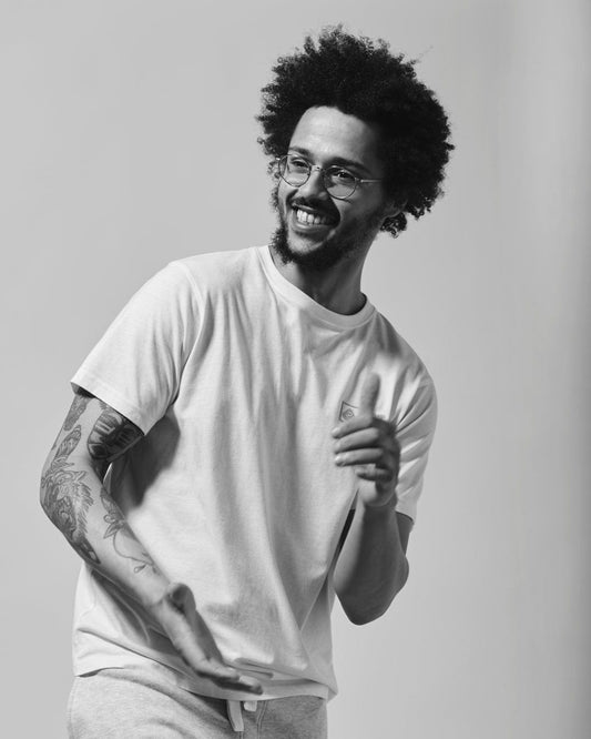 A black and white photograph of a smiling man with an afro hairstyle, wearing a casual Saltrock Original - Mens Short Sleeve T-Shirt - White with a crew neckline and shorts, showing tattoos on his arm, seemingly in mid-motion.