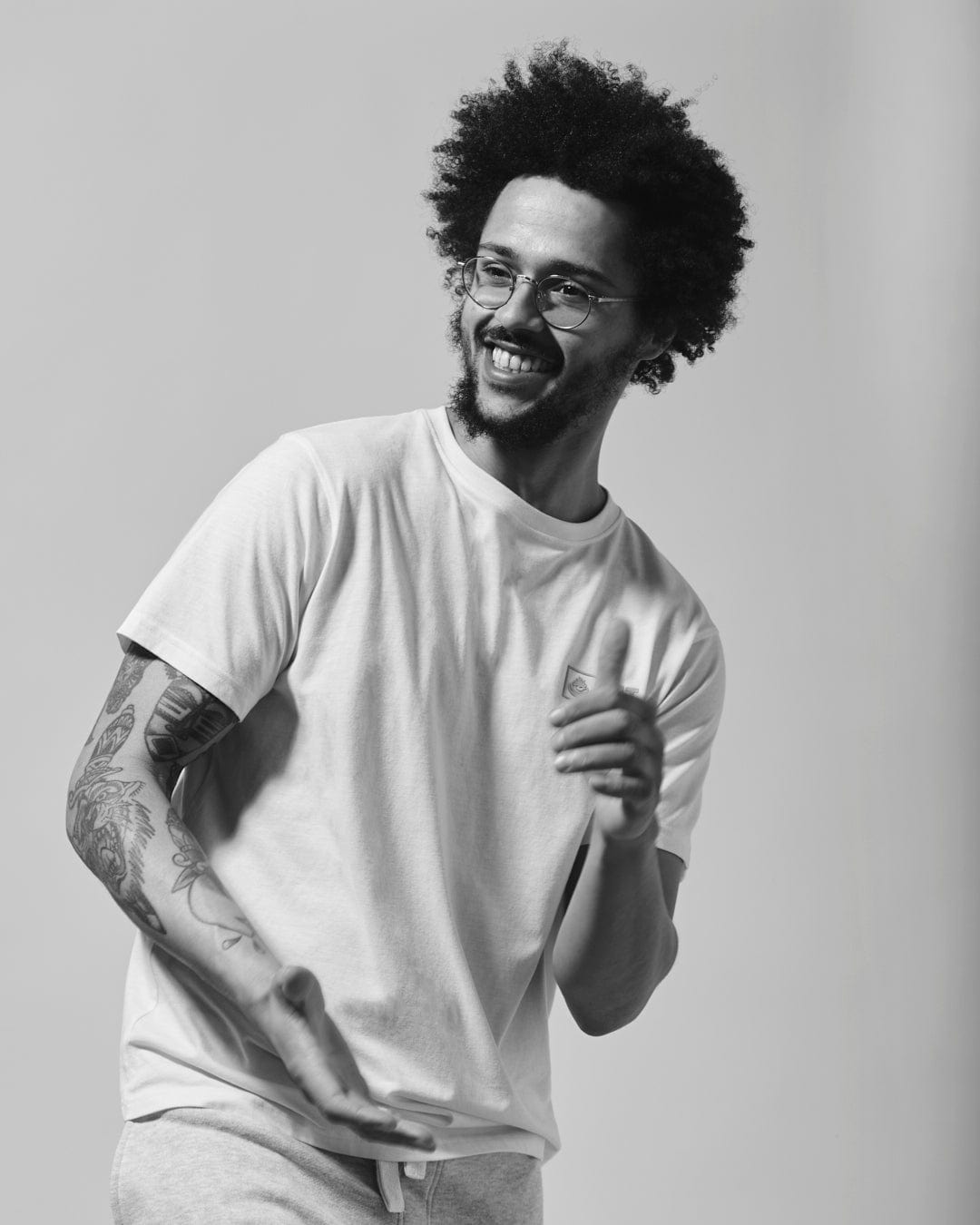A black and white photo of a smiling man with an afro hairstyle and a tattoo on his left arm, wearing a Saltrock Original - Mens Short Sleeve T-Shirt in Lilac made from lightweight material and sweatpants, appearing to be in mid m
