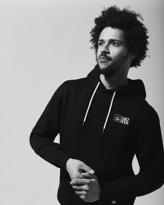 A person with curly hair wearing a Saltrock Original - Mens Pop Hoodie in Black, made of soft jersey material, stands against a light background, looking to the side with a thoughtful expression.