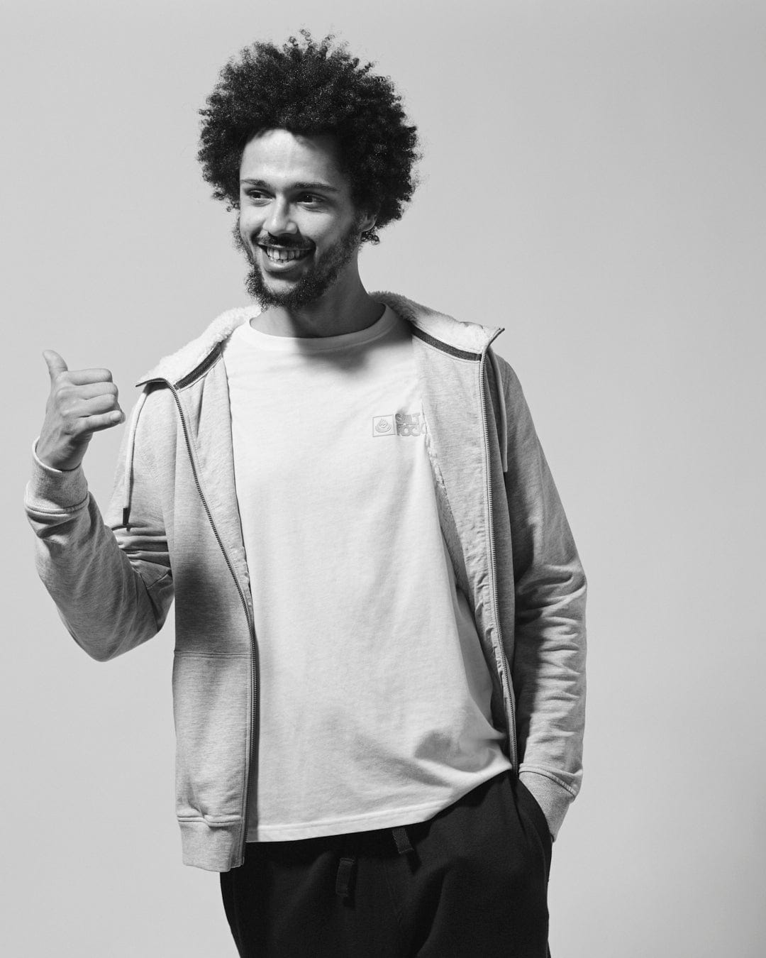A man with an afro hairstyle, smiling and giving a thumbs-up gesture to the camera, dressed in a casual hooded jacket featuring Saltrock branding over a Saltrock Original - Mens Short Sleeve T-Shirt - Dark Grey made of soft, lightweight material.