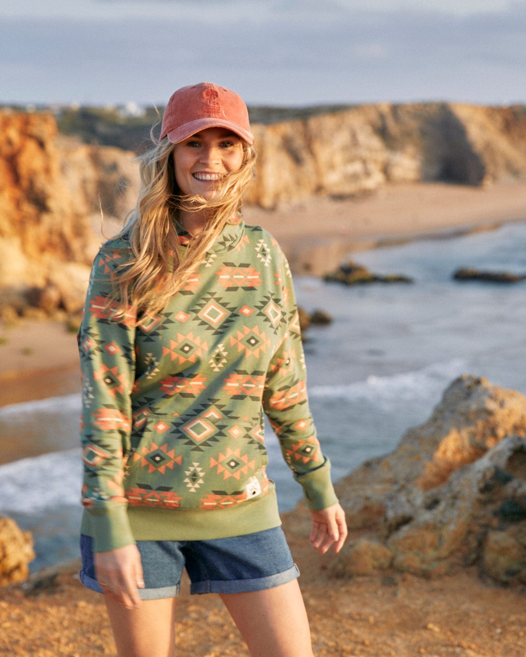 A woman wearing a Saltrock Aztec Santano - Womens Pop Hoodie - Green/Orange and cotton shorts on the beach.