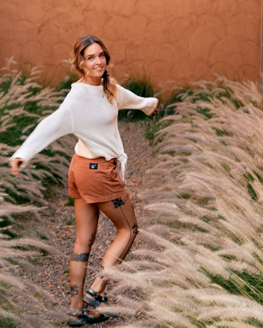 A woman with tattoos, wearing a Saltrock Beachcomber - Womens Tie Waist Jumper in Cream and brown shorts, playfully poses among tall grasses against a peach-colored wall.