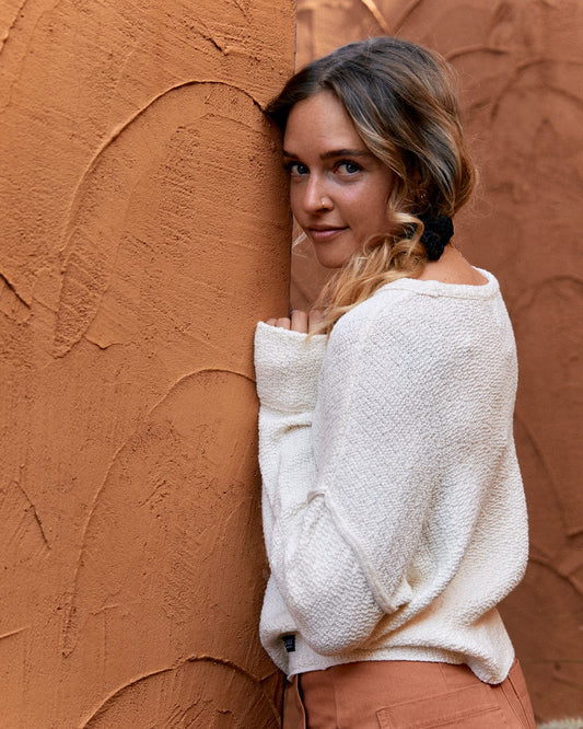 A woman with braided hair, wearing a Saltrock Beachcomber - Womens Tie Waist Jumper in Cream, smiles over her shoulder next to a textured terracotta wall.