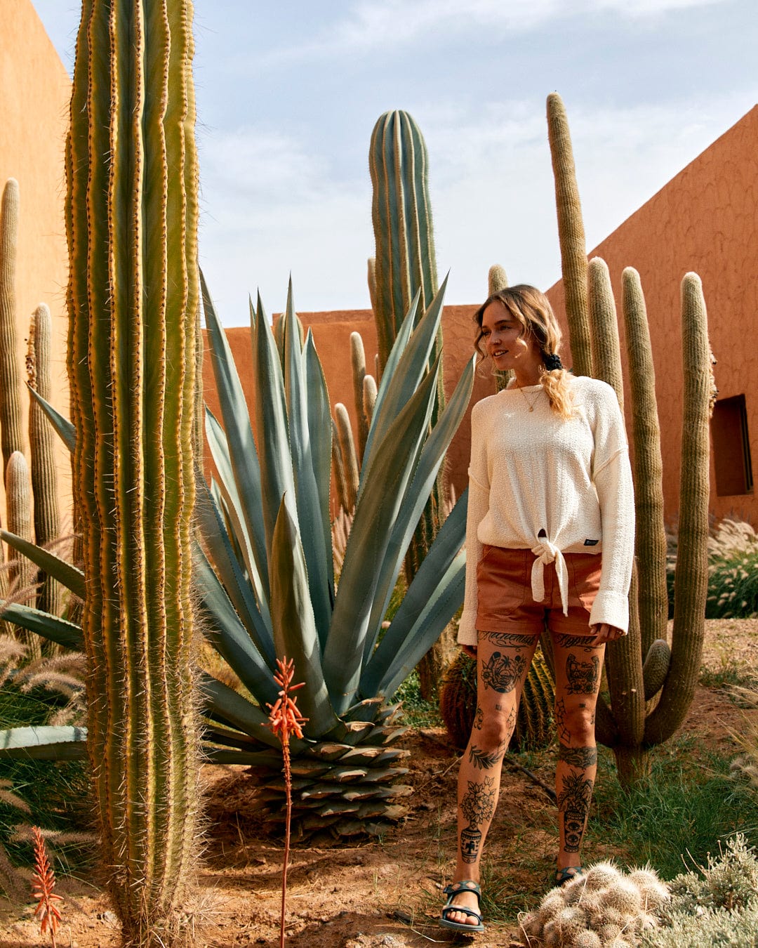 A woman in a Saltrock Beachcomber - Womens Tie Waist Jumper in Cream and patterned leggings smiles while standing near tall cacti in a desert garden.