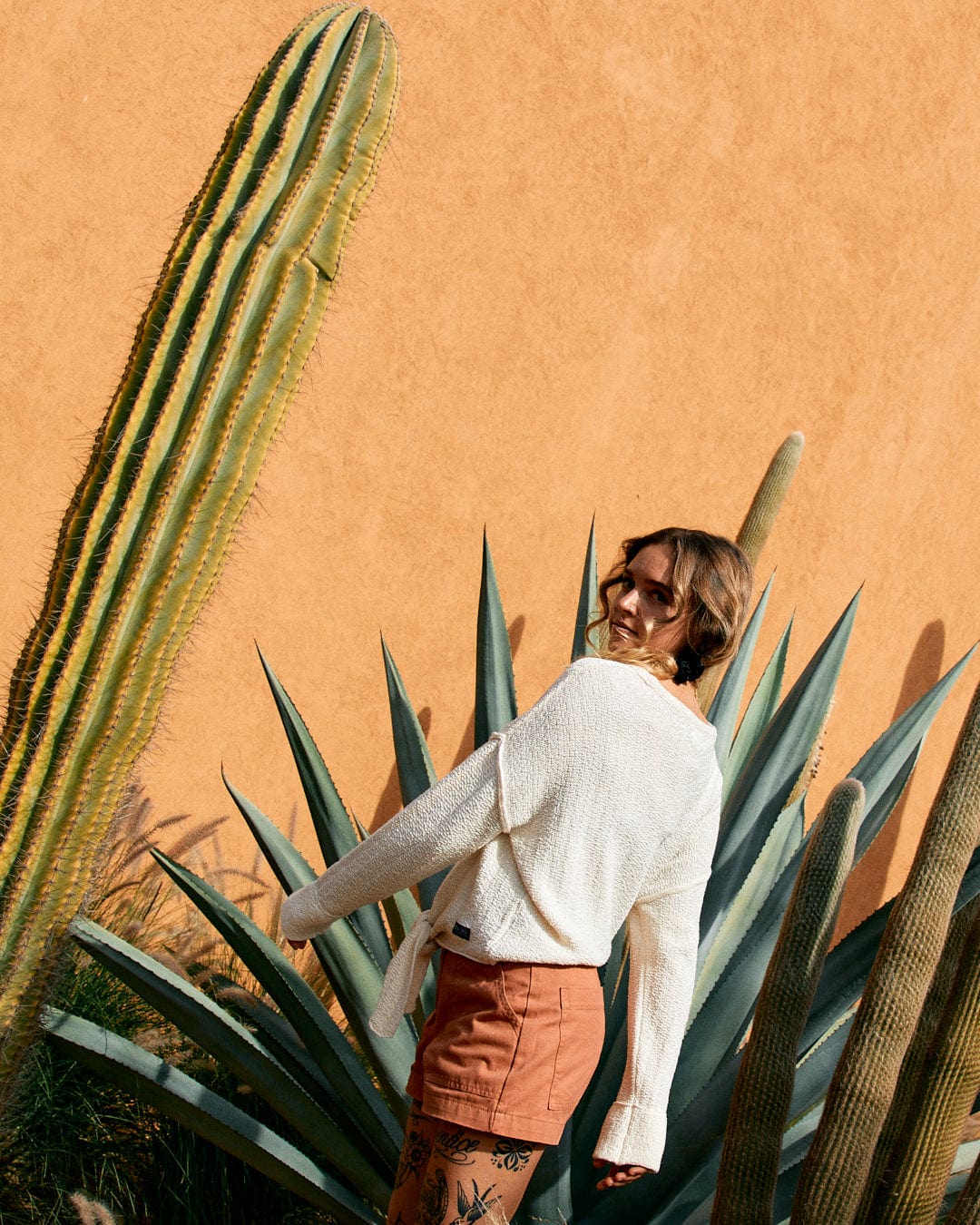 A woman looking over her shoulder near tall cacti and agave plants, against an orange textured wall, wearing a Saltrock Beachcomber - Womens Tie Waist Jumper in Cream and terracotta tie-waist shorts.