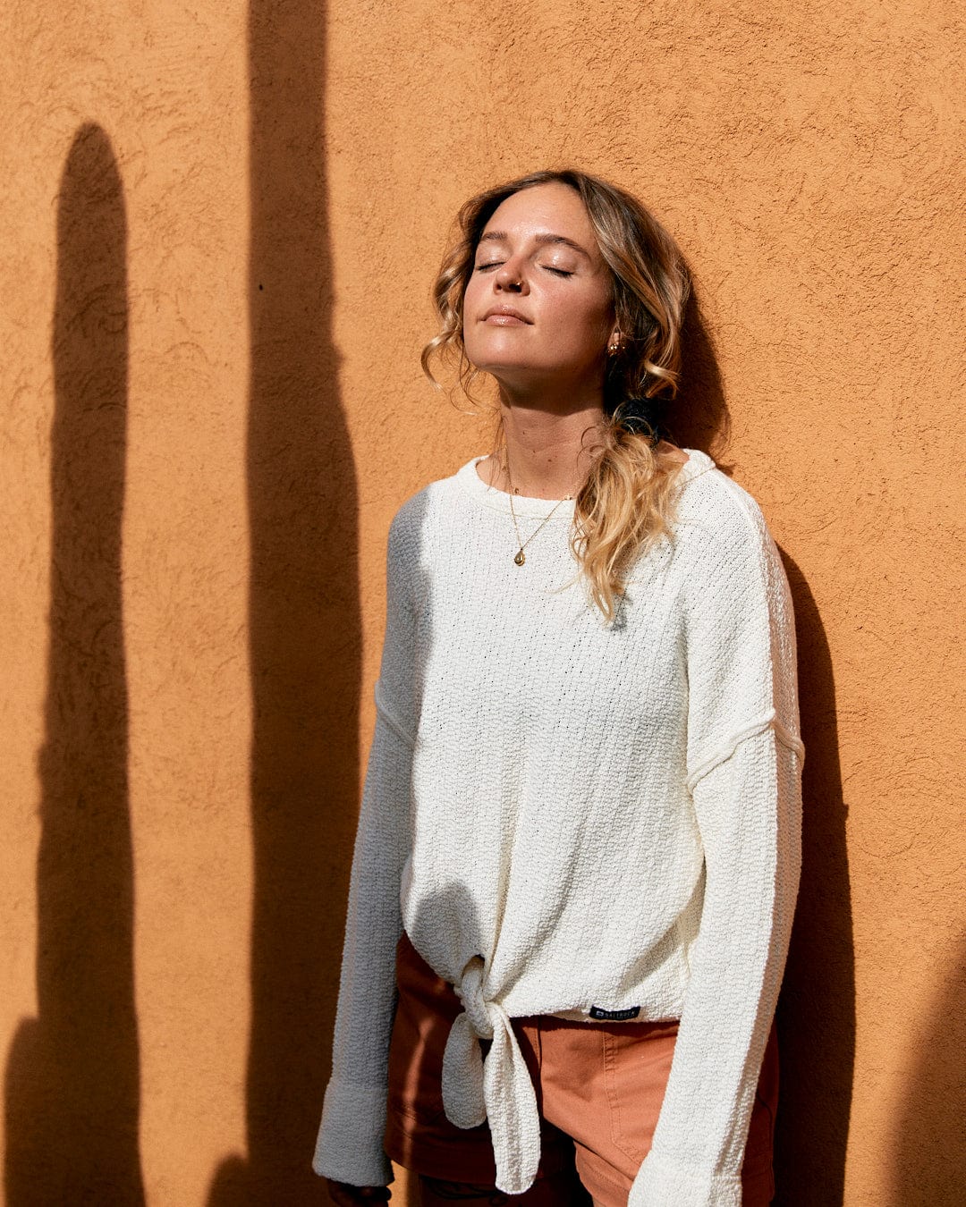 A woman with closed eyes enjoys the sunlight against a warm ochre wall, casting a long shadow beside her. She wears a casual white textured knit sweater and rust-colored pants, such as the Beachcomber - Womens Tie Waist Jumper in Cream from Saltrock.