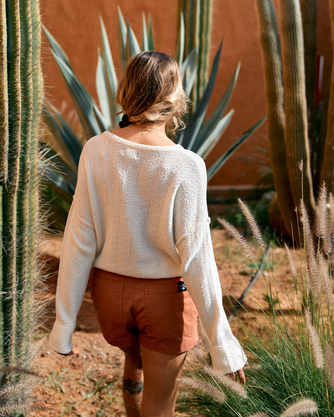 A woman in a Saltrock Beachcomber - Womens Tie Waist Jumper in Cream and brown shorts standing in front of tall cacti, viewed from behind.