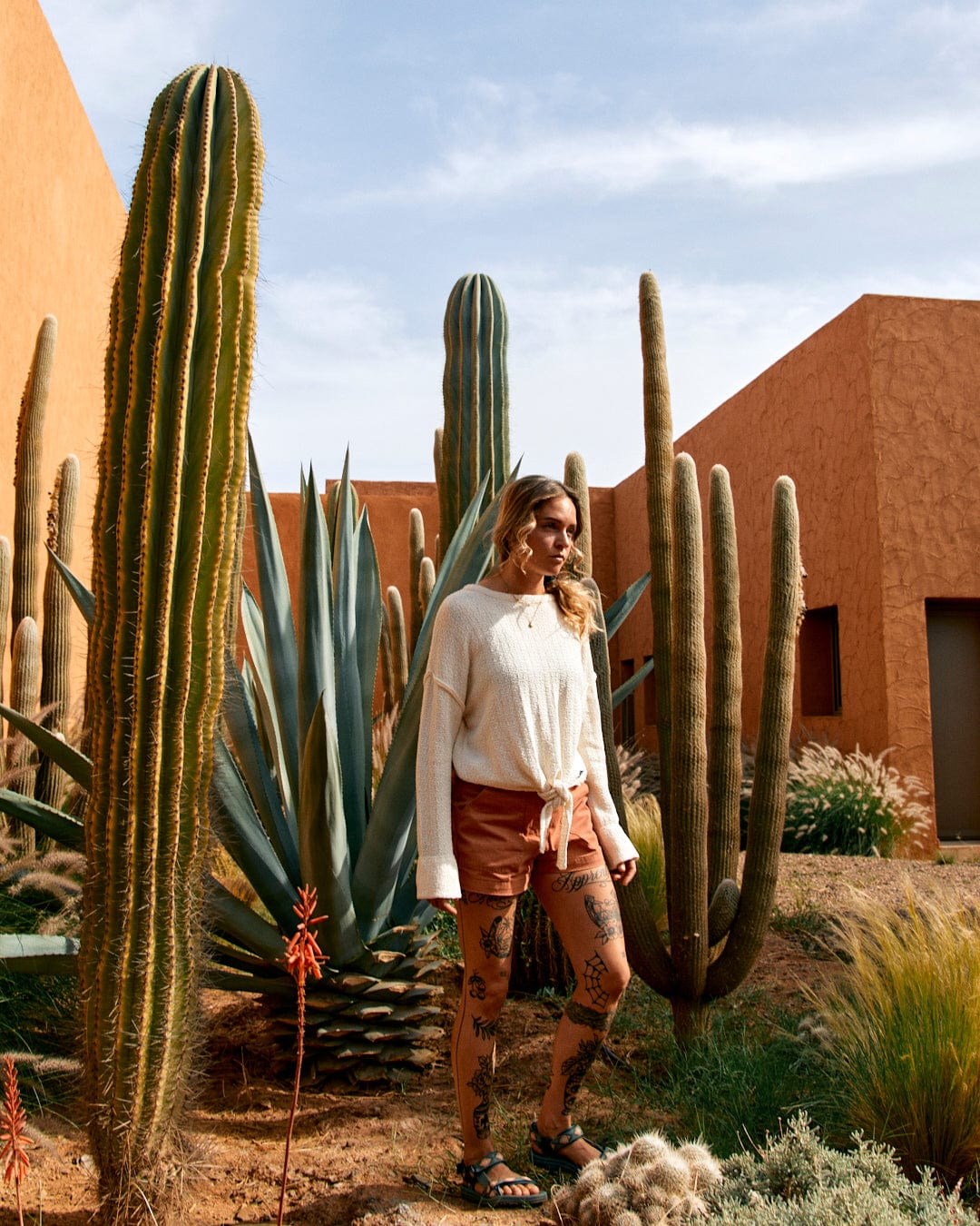A woman stands amid large cacti, wearing a Saltrock Beachcomber - Womens Tie Waist Jumper in Cream, and brown shorts with a tie waist, featuring visible tattoos on her legs.