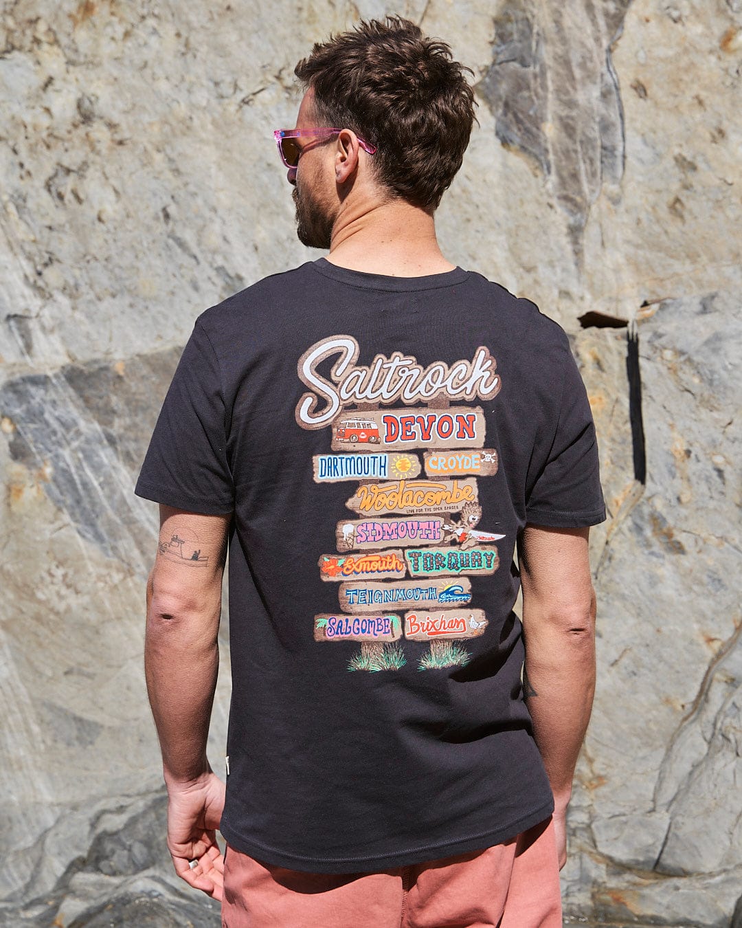 The back of a man wearing Saltrock sunglasses and a Beach Sign Devon - Mens T-Shirt - Dark Grey.