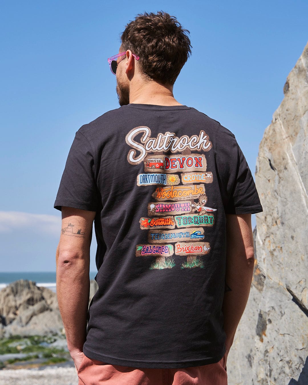 A man wearing Saltrock sunglasses and a Saltrock Beach Sign Devon - Mens T-Shirt - Dark Grey looking at the ocean.