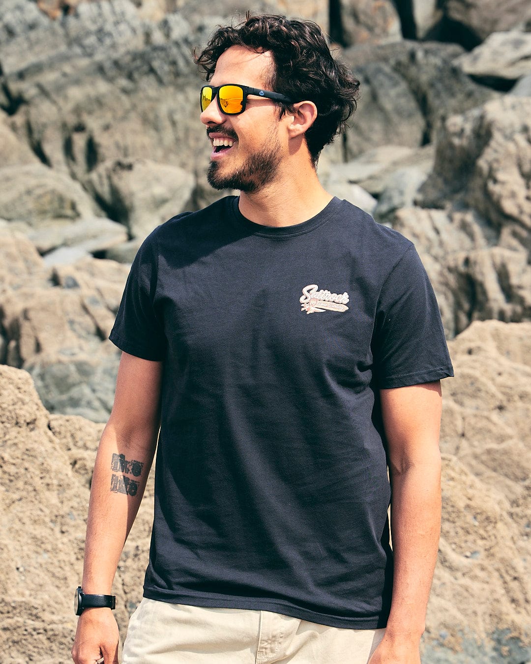 A man wearing sunglasses and a Saltrock Beach Sign Wales - Mens - Short Sleeve T- Shirt - Dark Grey in front of rocks.