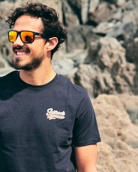 A man wearing sunglasses and a Saltrock Beach Sign Wales - Mens - Short Sleeve T-Shirt - Dark Grey in front of rocks.