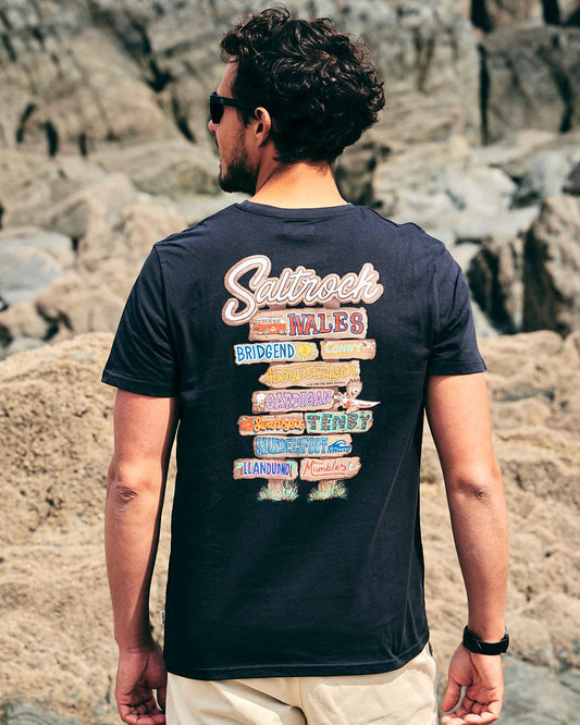 A man wearing a Saltrock Beach Sign Wales - Mens - Short Sleeve T- Shirt - Dark Grey looking at the rocks.