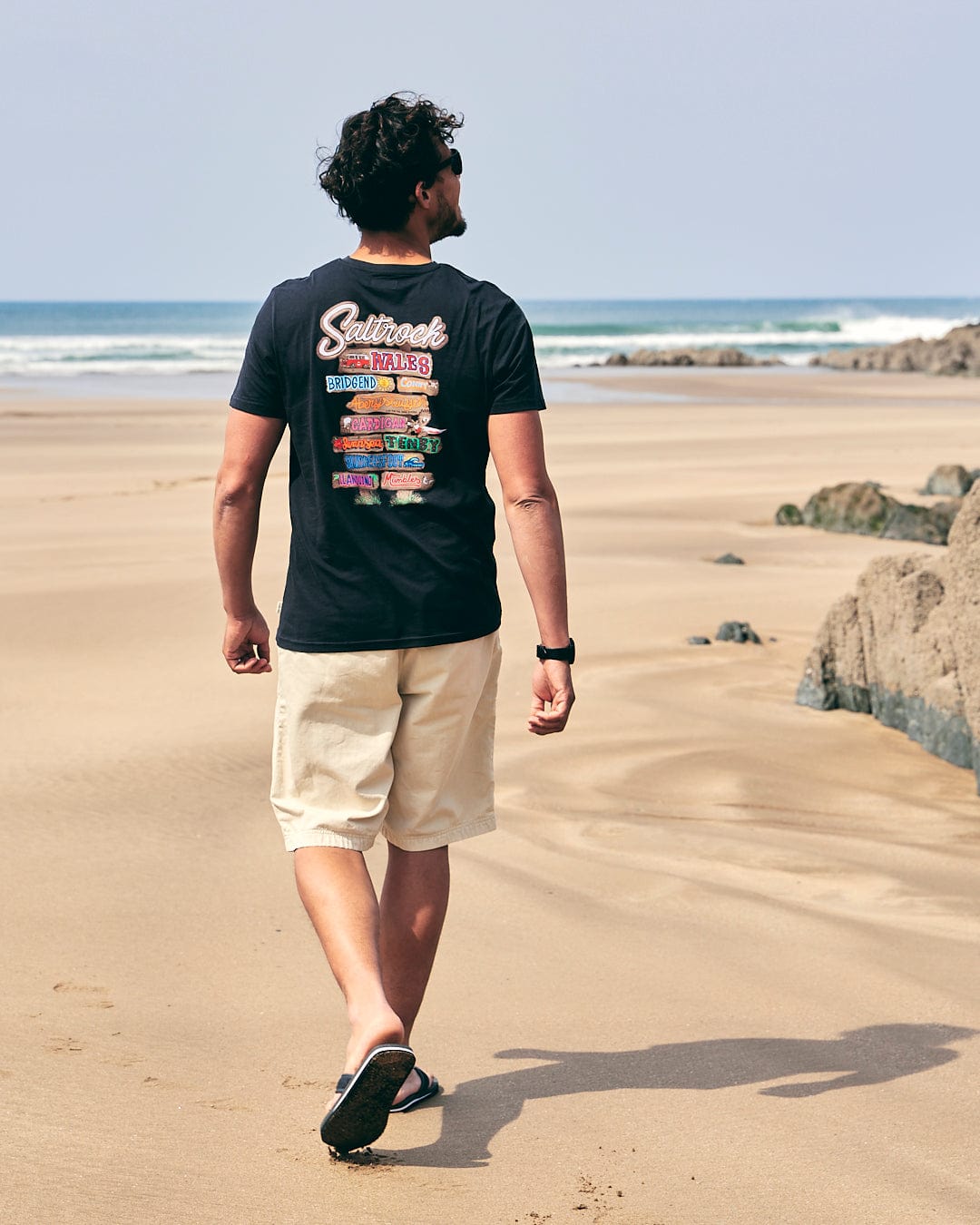 A man walking on the beach wearing a Saltrock - Beach Sign Wales - Mens - Short Sleeve T-Shirt - Dark Grey.