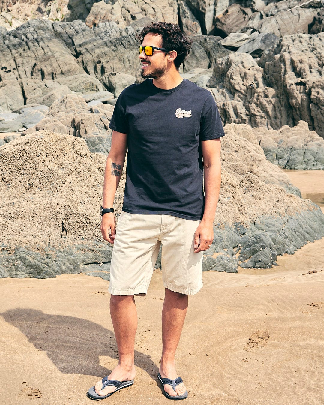 A man standing on a beach with a Saltrock - Beach Sign Wales - Mens - Short Sleeve T-Shirt - Dark Grey surfboard.