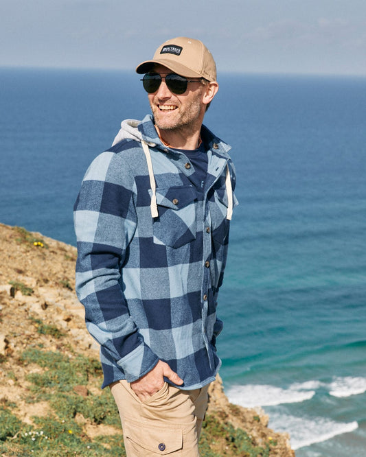 Man in a Saltrock Beale - Mens Hooded Long Sleeve Shirt - Blue with a detachable hood and cap smiling by the sea.
