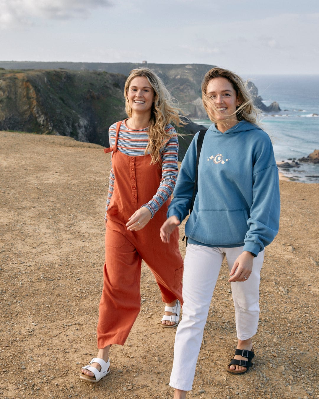 Two women walking outdoors near a coastal cliffside, dressed in casual attire, one in a red jumpsuit and a striped shirt embellished with floral patterns, the other in white pants and a Better Days - Womens Pop Hoodie - Blue from Saltrock.