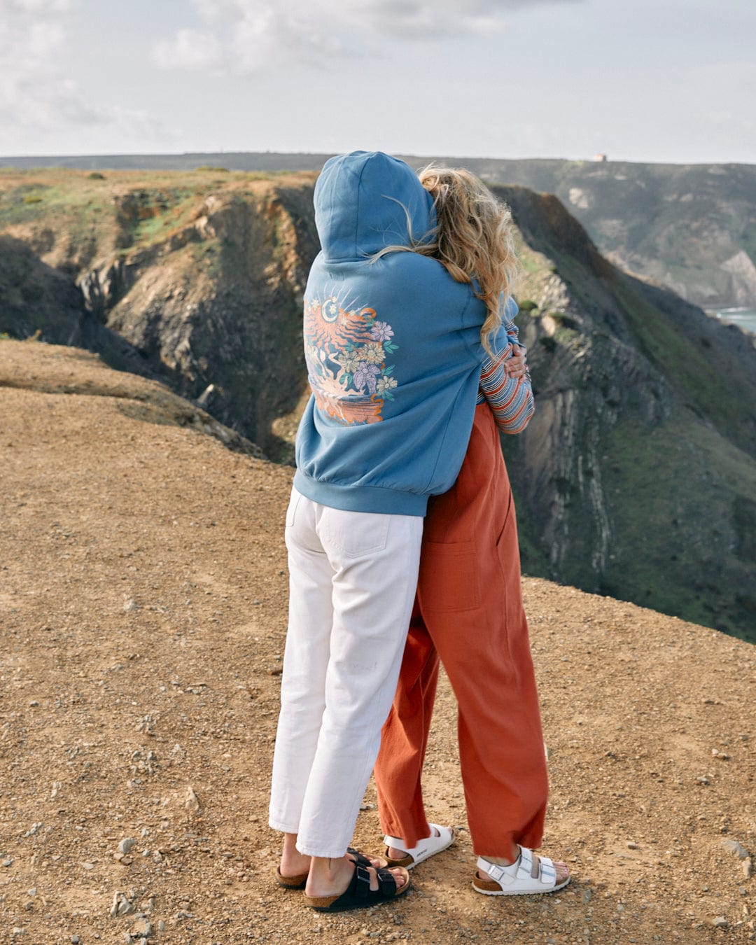 Two people embrace while standing on a cliff overlooking a scenic coastal view; one wears a Saltrock Better Days - Womens Pop Hoodie in Blue with a floral design on the back, and the other a pair of red trousers and a white top.
