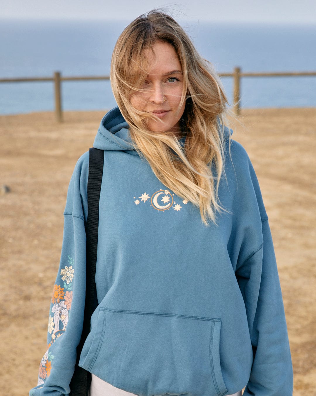 A woman wearing a Saltrock Better Days - Womens Pop Hoodie in Blue with brighter floral patterns on the sleeves stands outdoors with a sandy beach in the background.