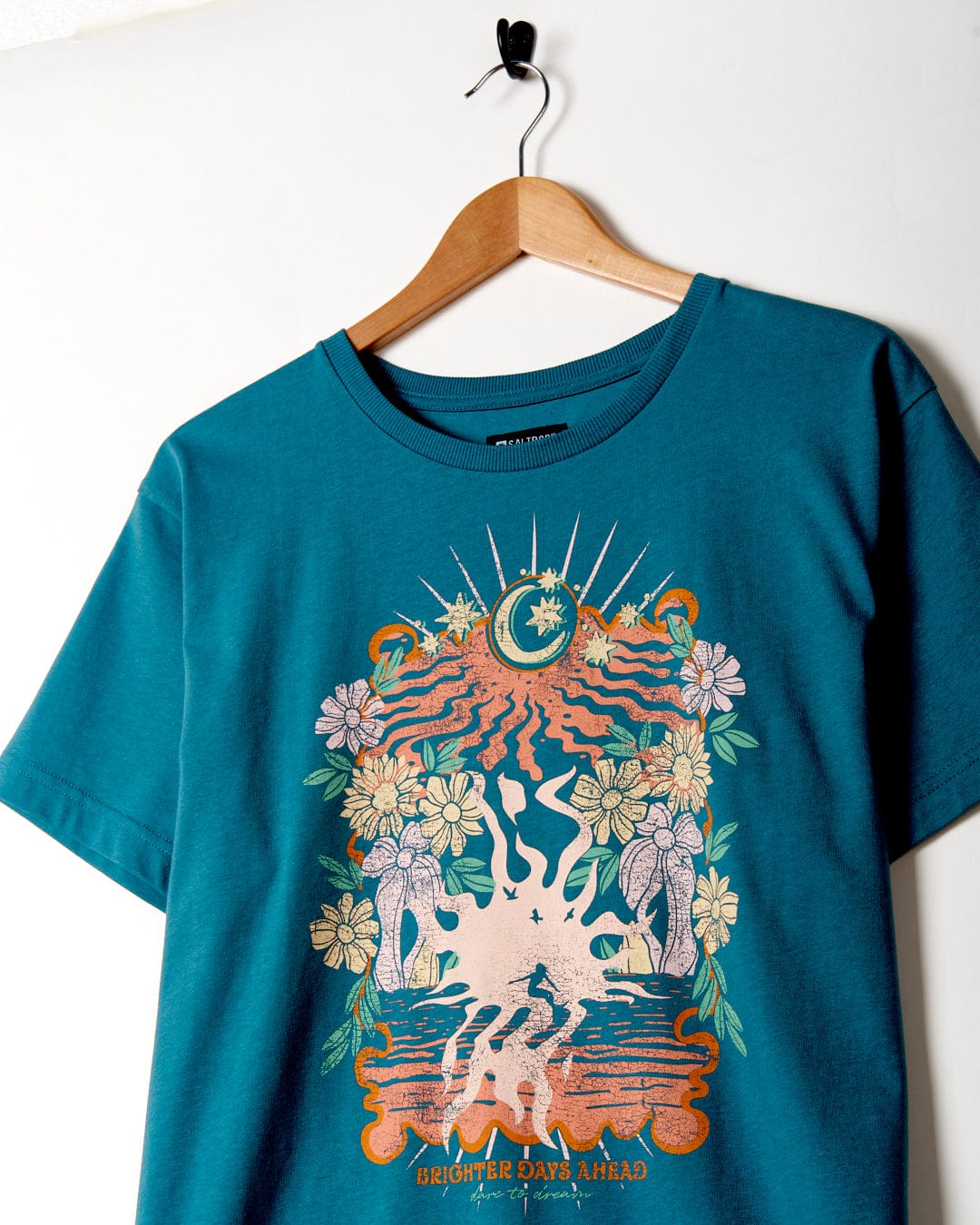 Better Days - Recycled Womens Short Sleeve Relaxed T-Shirt in Teal by Saltrock, hanging on a wall-mounted hook, featuring a sun, moon, and floral design with the text "brighter days ahead.