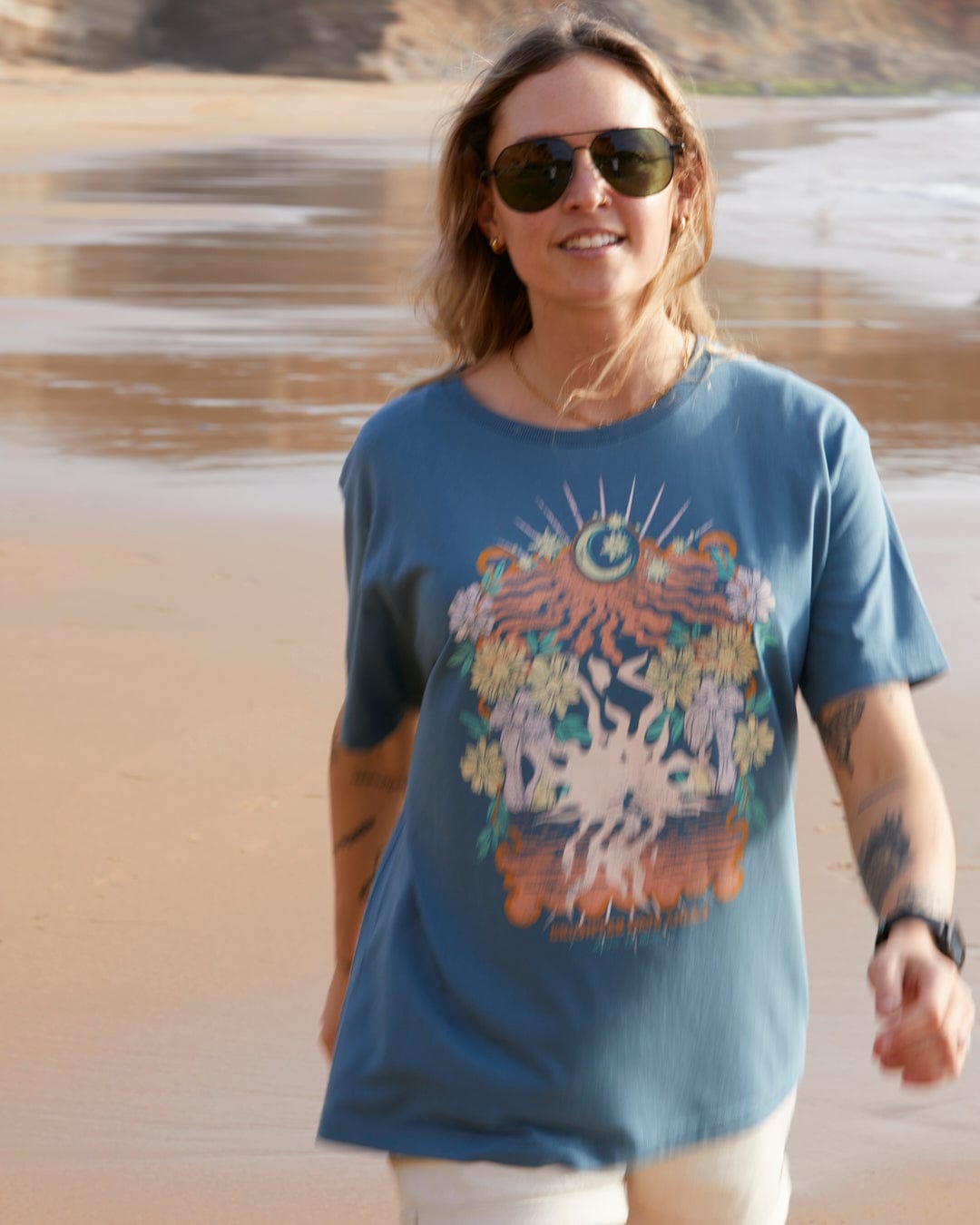 A woman in sunglasses and a relaxed loose fit Better Days - Recycled Womens Short Sleeve Relaxed T-Shirt in Teal walks on a sandy beach, with the background slightly blurred.
