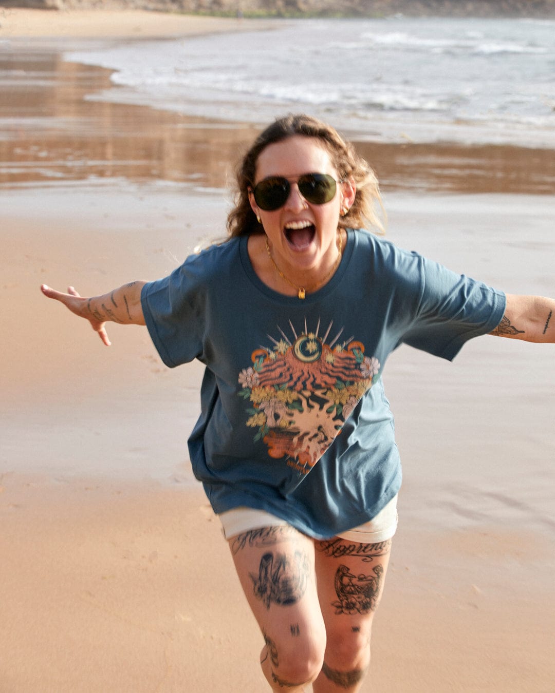 A joyful woman with tattoos wearing sunglasses and a Saltrock Better Days - Recycled Womens Short Sleeve Relaxed T-Shirt in Teal runs on a sandy beach, her arms outstretched.