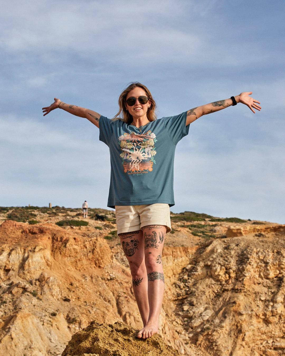 Woman standing on a rocky hill with arms spread wide, smiling, wearing a Saltrock Better Days - Recycled Womens Short Sleeve Relaxed T-Shirt in Teal and shorts, with a cloudy sky background and another person in the distance.