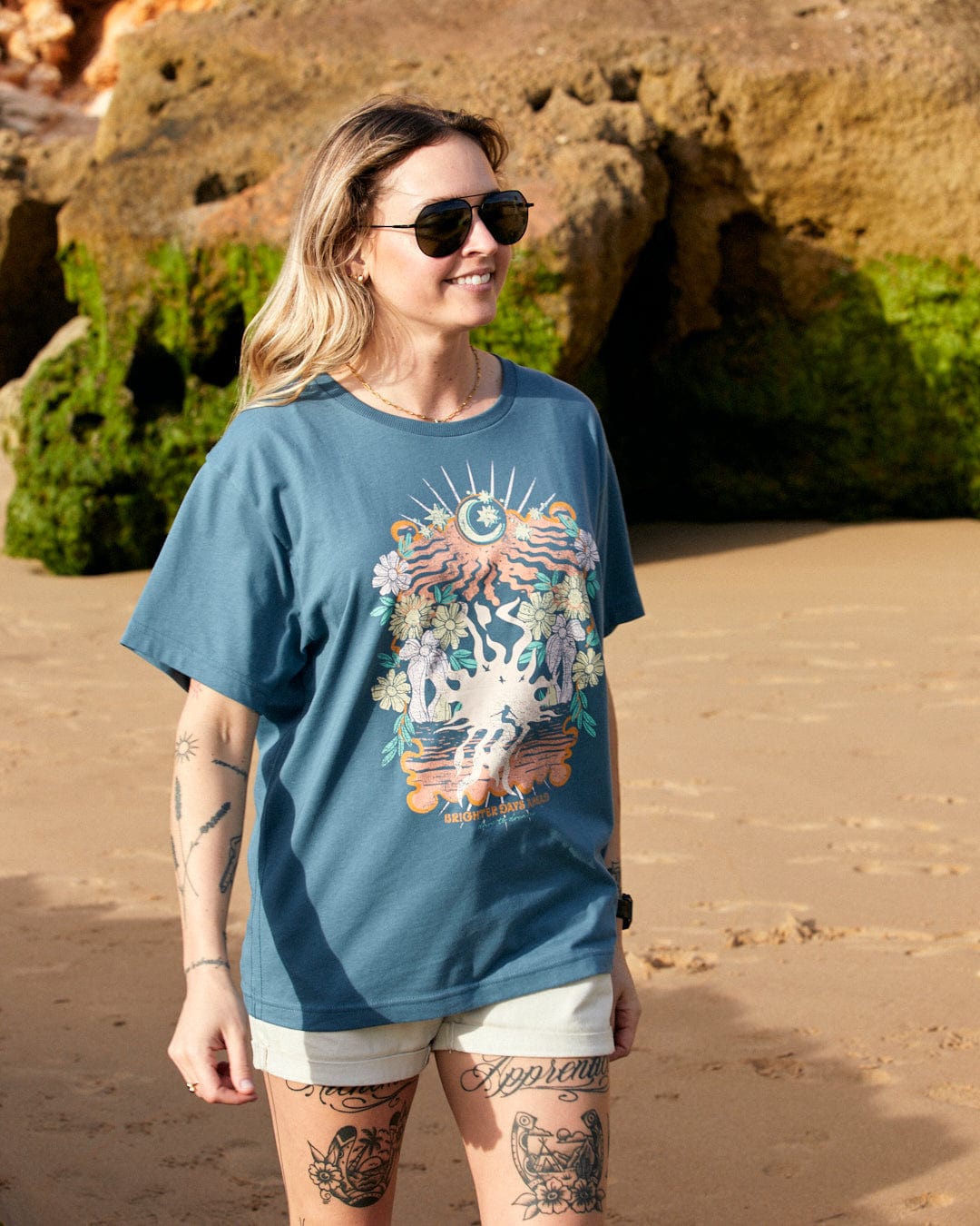 A woman wearing sunglasses and a Better Days - Recycled Womens Short Sleeve Relaxed T-Shirt in Teal made by Saltrock stands on a sandy beach with rocky cliffs in the background.