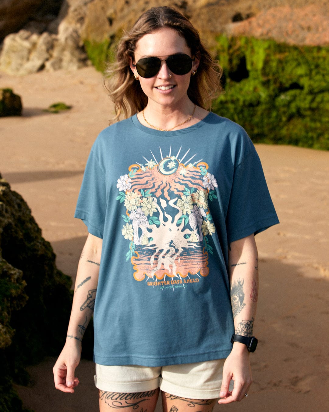 A woman in sunglasses stands on a sandy beach, wearing a blue Saltrock t-shirt with a colorful floral surfing luna graphic and denim shorts.