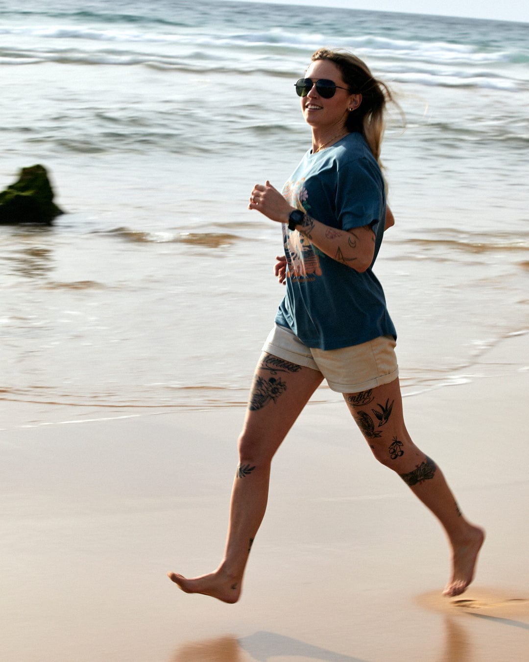 A woman with tattoos running joyfully on a beach, wearing a Saltrock Better Days - Recycled Womens Short Sleeve Relaxed T-Shirt in Teal and shorts, with sunglasses on a sunny day.