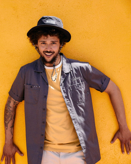A man with curly hair wearing a hat and a short-sleeved shirt smiles, leaning against a bright yellow wall. His shirt, the Saltrock Boulevard - Mens Short Utility Sleeve Shirt - Dark Grey, crafted in utility-style cotton canvas, features chest pockets and embroidered Saltrock branding.