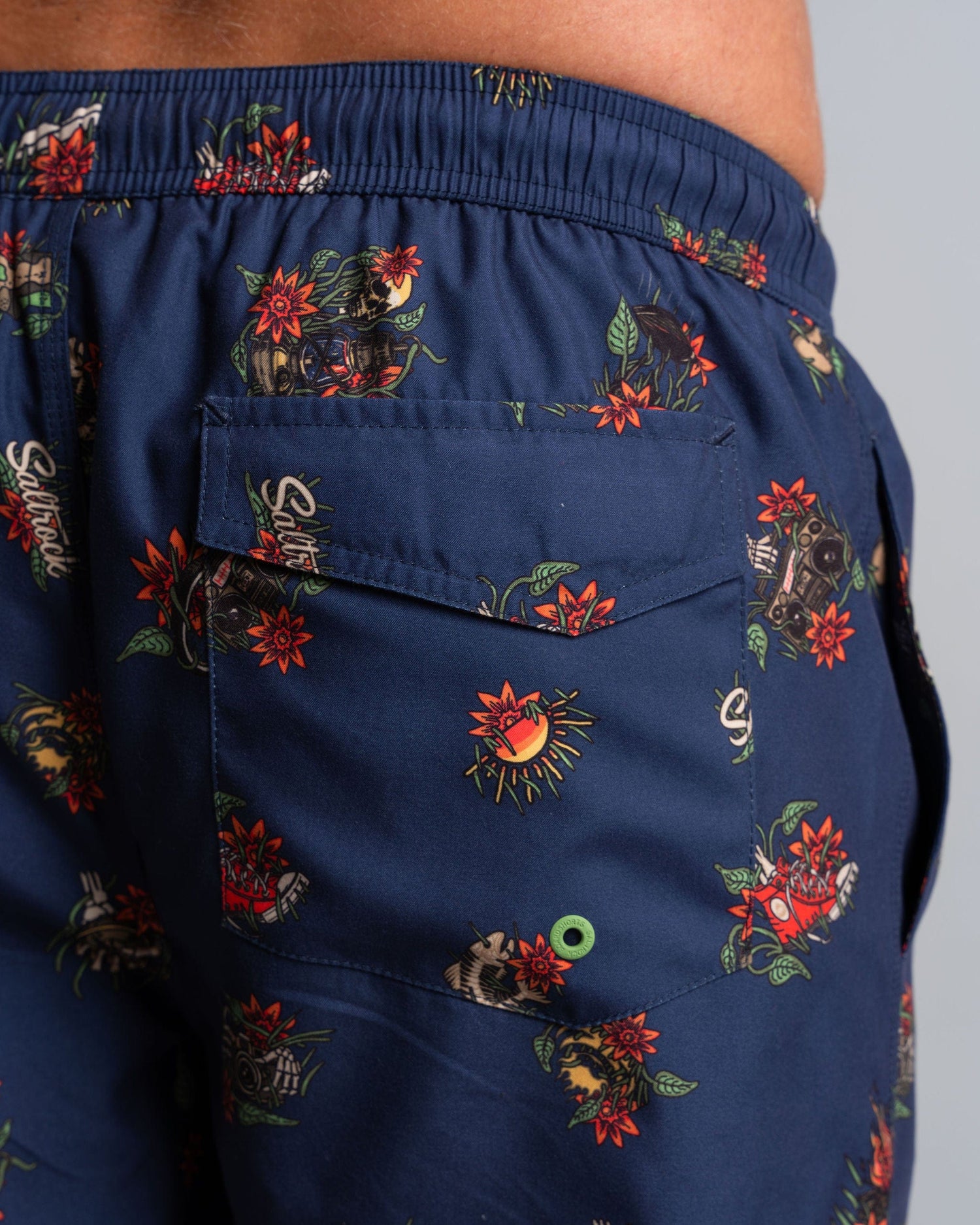 Close-up of a person wearing Saltrock's Wanderer men's swim shorts in blue, showcasing the brand's vibrant floral and skull design. The mesh-lined shorts feature an elasticated waist and a back pocket with a small green grommet.