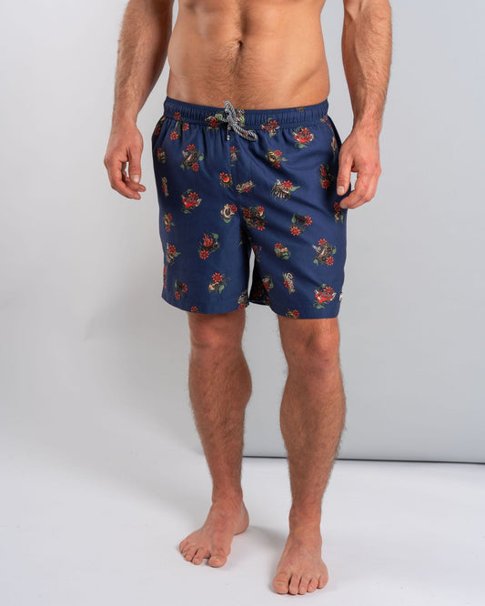 A man stands barefoot against a plain backdrop, wearing Saltrock's "Wanderer" men's blue swim shorts with a floral design and elasticated waist, featuring a mesh-lined interior for comfort, confidently displaying his shirtless torso.