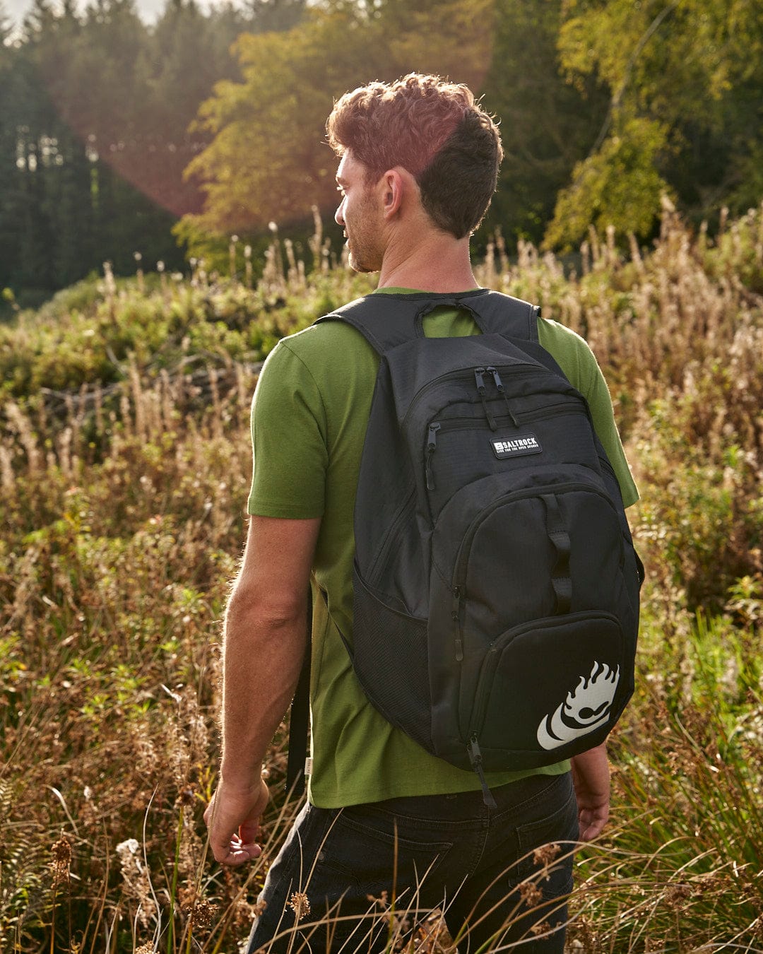A man walking through a field with a Saltrock Boardwalk - Backpack - Black, made of water-resistant ripstop fabric and equipped with a cooler pocket.