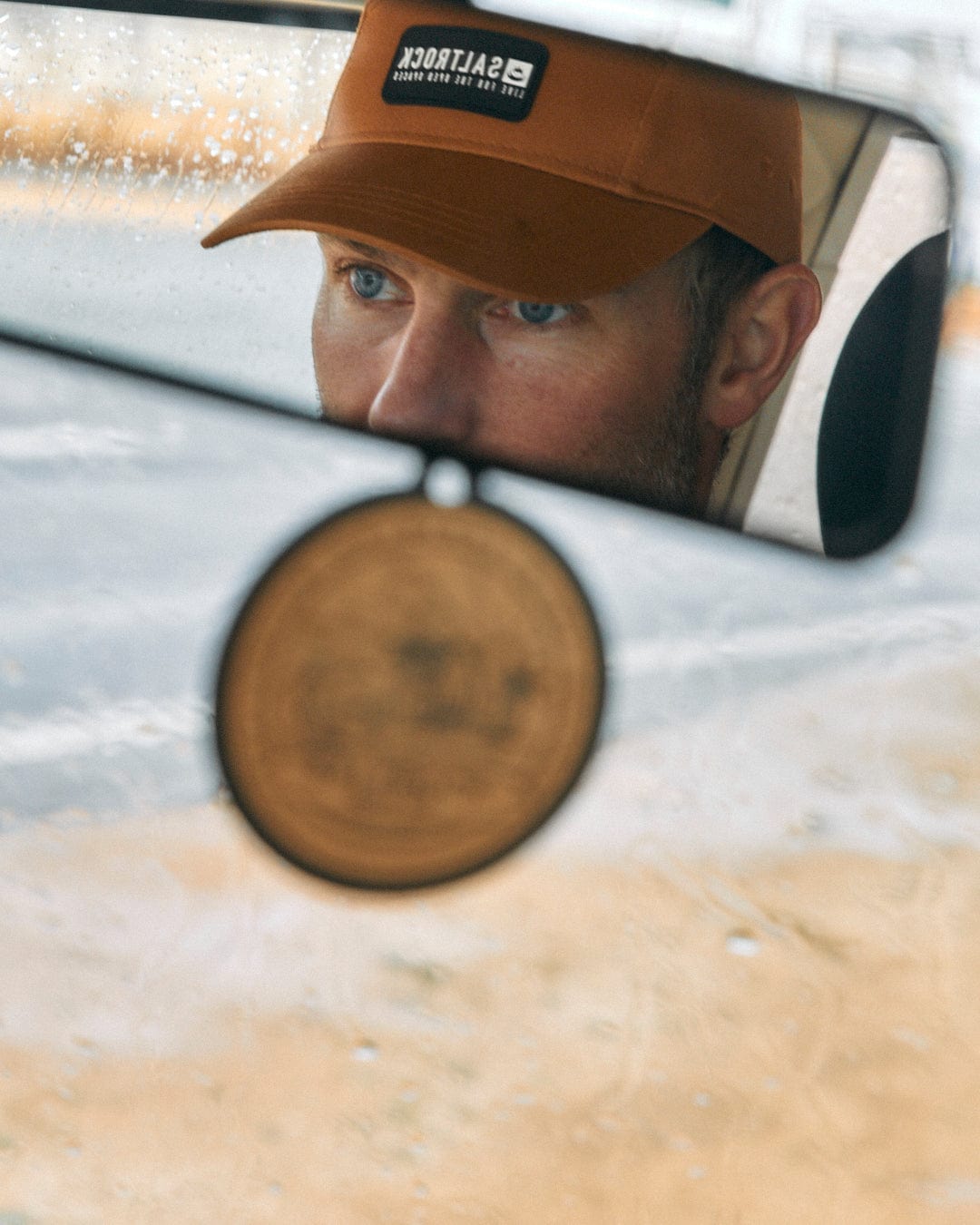 Man with blue eyes seen in car’s rear-view mirror, wearing a brown Saltrock Boardwalk - 5 Panel Cap, with raindrops visible on the window.