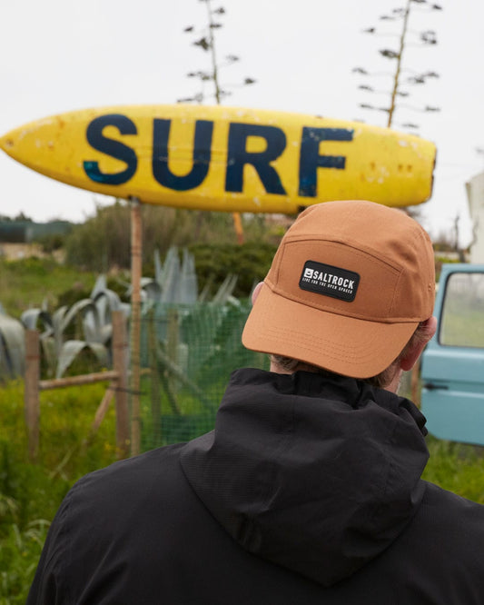 Person wearing a black jacket and an orange cap with a curved visor, viewed from behind, looking at a yellow surfboard with "surf" written on it, positioned against a rural background.