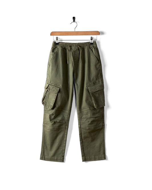 The Saltrock Bora Bora Kids Chino Trouser in dark green, featuring a baggy fit, cargo-style pockets, and an elasticated waist, hangs on a wooden hanger against a plain white background.