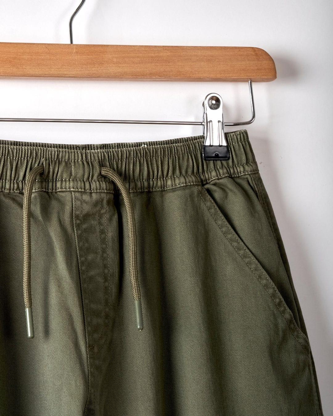 Close-up of Saltrock's Bora Bora Kids Chino Trouser in dark green, featuring a baggy fit and elastic waistband, displayed on a wooden hanger against a white background.