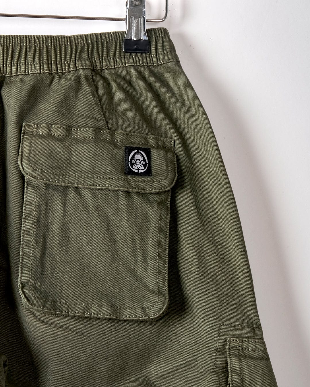 Close-up of Saltrock Bora Bora Kids Chino Trousers in dark green, featuring an elasticated waist, flap cargo-style pockets, and a small black and white patch with a face logo.