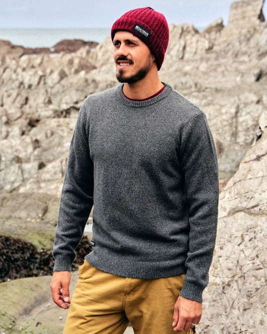 A man wearing a Saltrock Bowen - Mens Knitted Crew - Dark Grey sweater and a beanie standing on rocks.