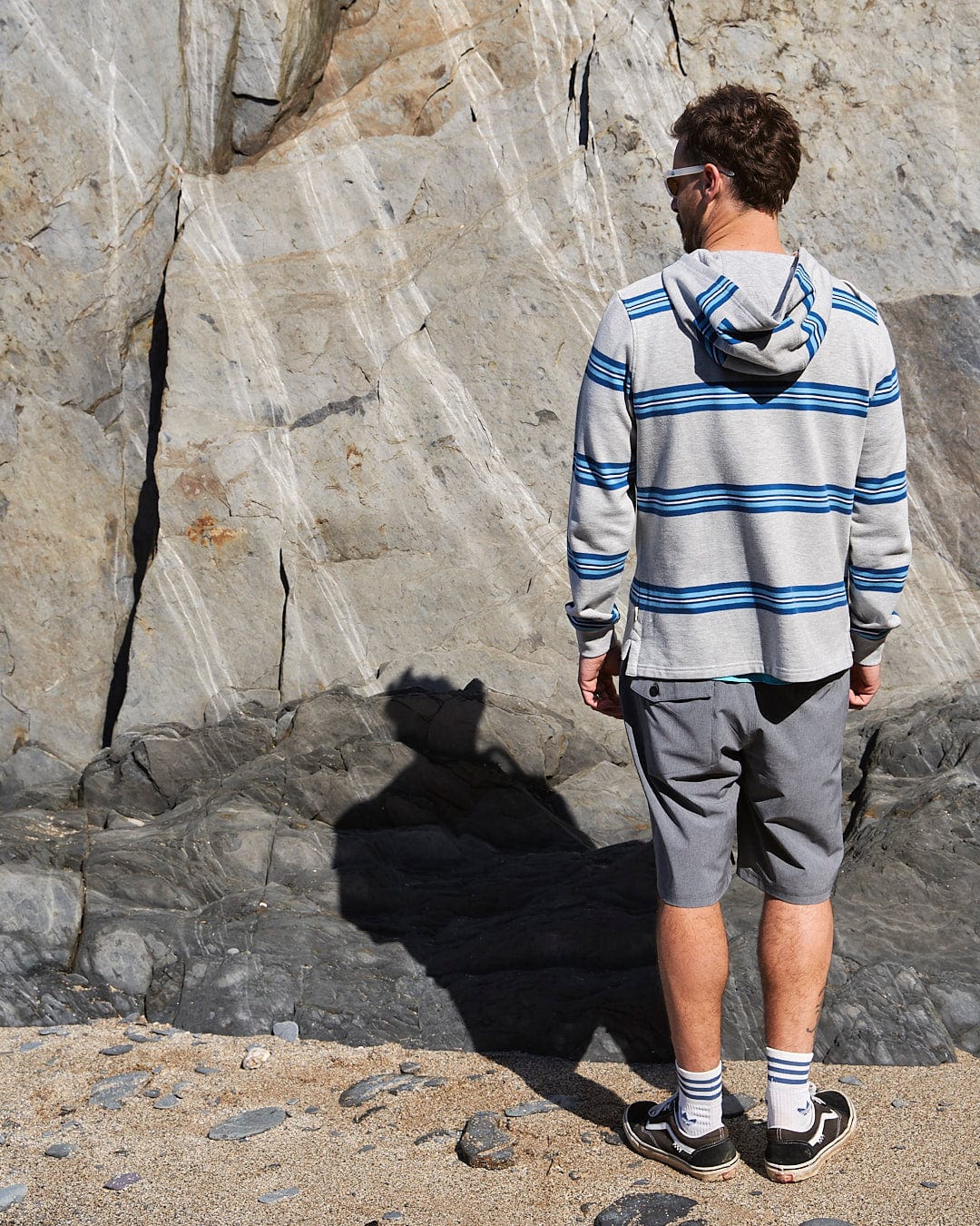 A man wearing the Saltrock Byron - Mens Pop Hoodie - Grey standing next to a rock.