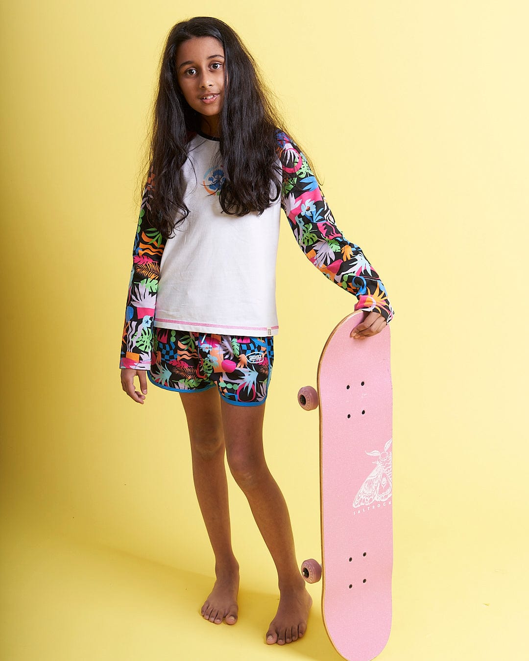 A girl holding a Saltrock Zephyr - Kids Boardshort - Blue skateboard in front of a yellow background.