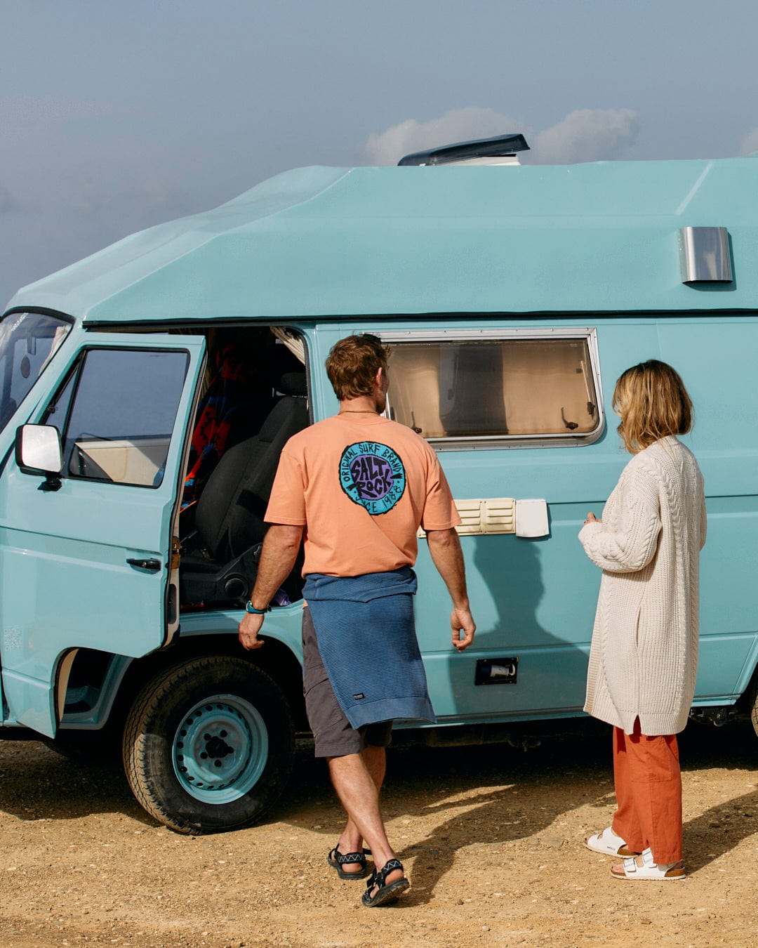 Two individuals standing beside a turquoise van on a dusty terrain, one wearing a Saltrock Womens Knitted Longline Cardigan in Cream.