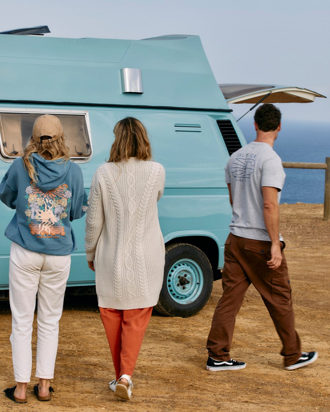 Three people, one wearing a Saltrock Cable - Womens Knitted Longline Cardigan in Cream, walking towards a blue camper van by the seaside.