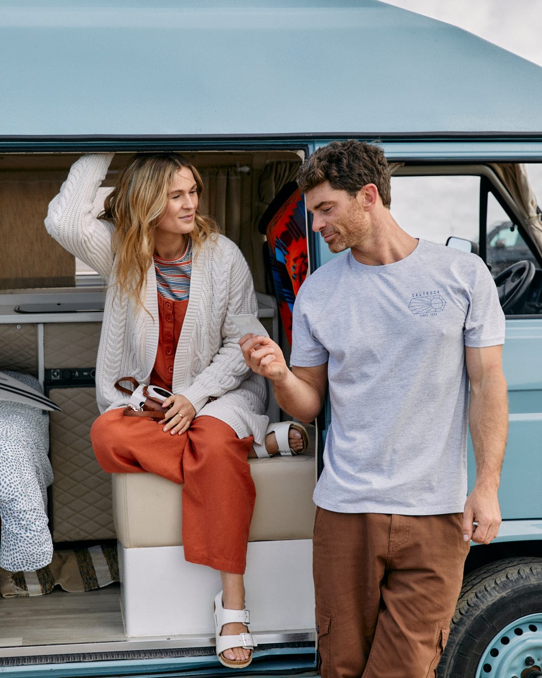 A man and a woman, the latter cozily wrapped in a Saltrock Cable - Womens Knitted Longline Cardigan in Cream, enjoying a moment together by a camper van.